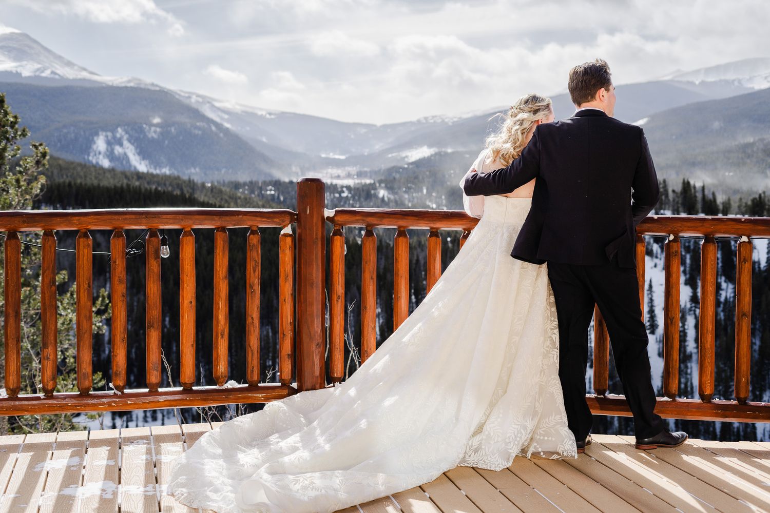 Bride and groom standing on a wooden deck overlooking snow-capped mountains in winter scenery.