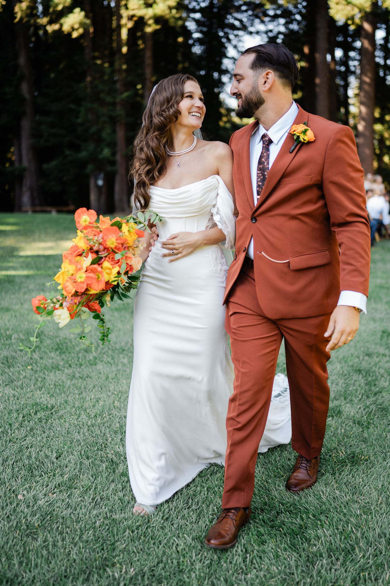 Smiling bride and groom walking together, groom in rust suit, bride holding vibrant orange bouquet.