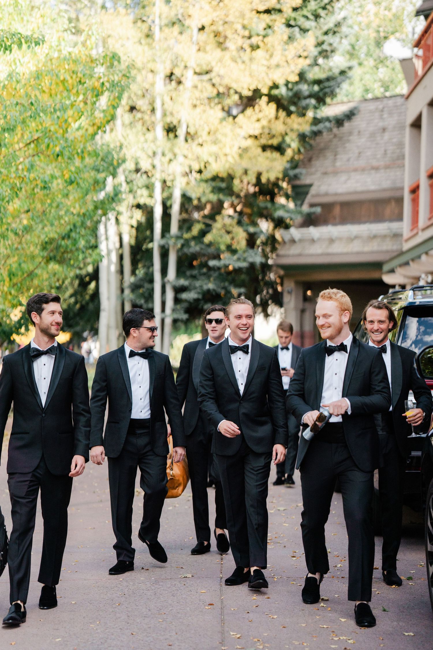 Groomsmen in black tuxedos and bow ties walking together outdoors among autumn trees.