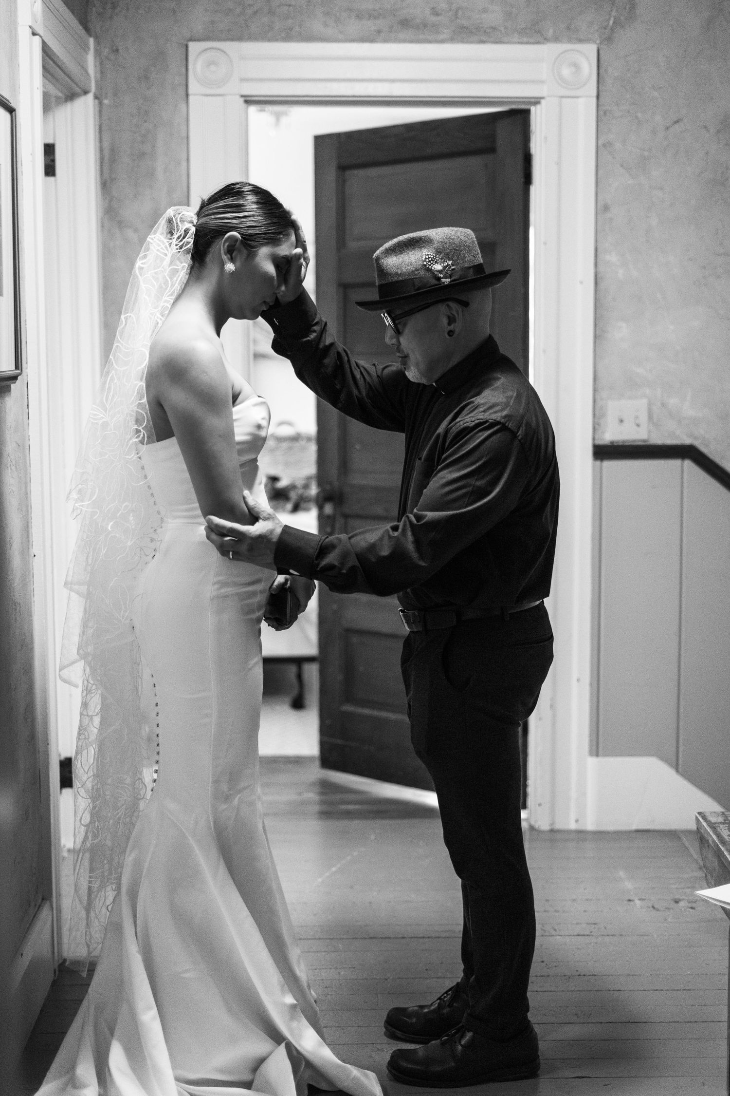 Black and white photo of groom tenderly wiping bride's tears in hallway before wedding ceremony.
