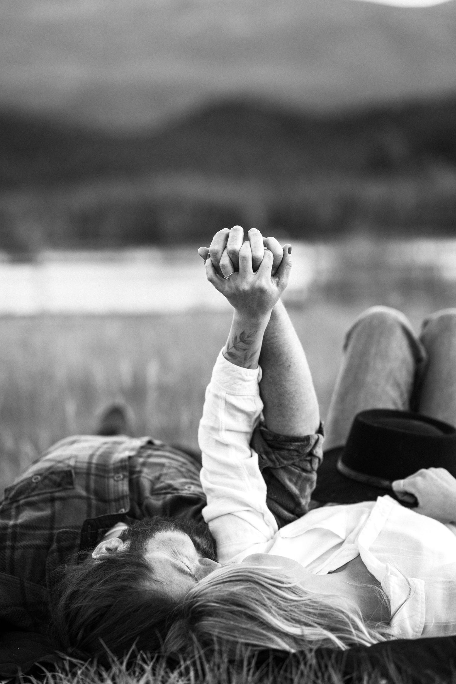 Black and white photo of couple lying in grass holding hands raised together near a mountain lake.