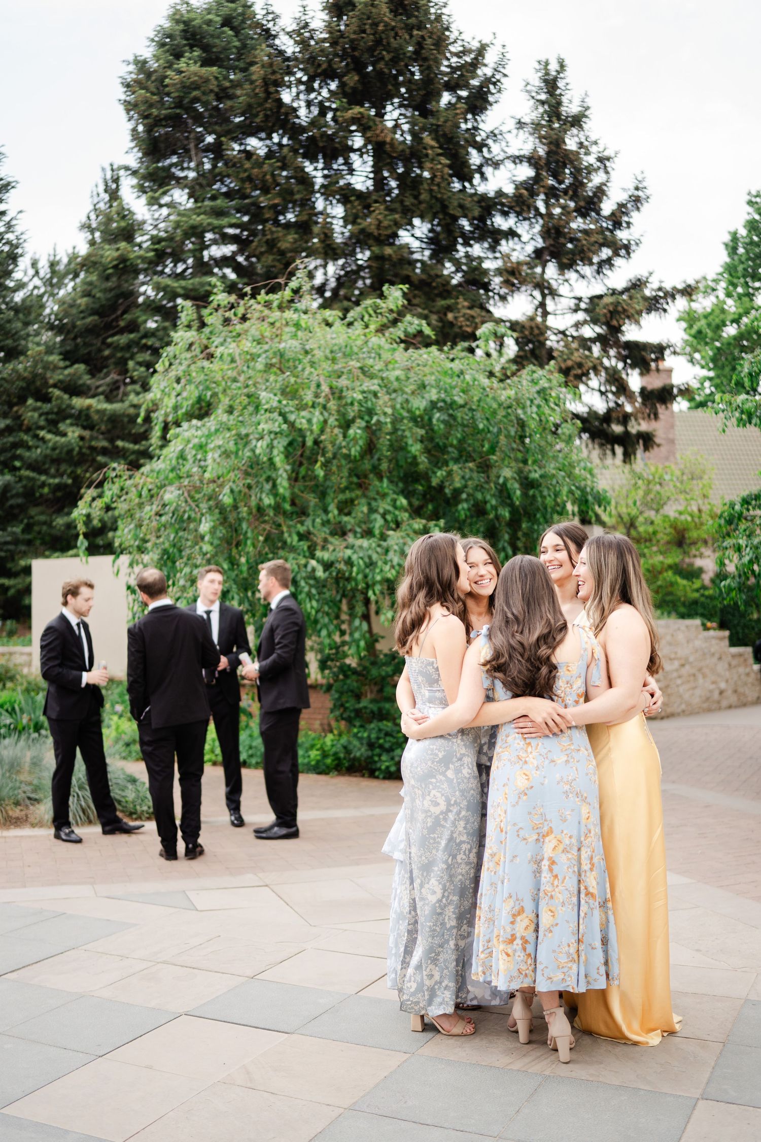 Bridesmaids in floral dresses embracing at outdoor wedding reception with groomsmen in background.