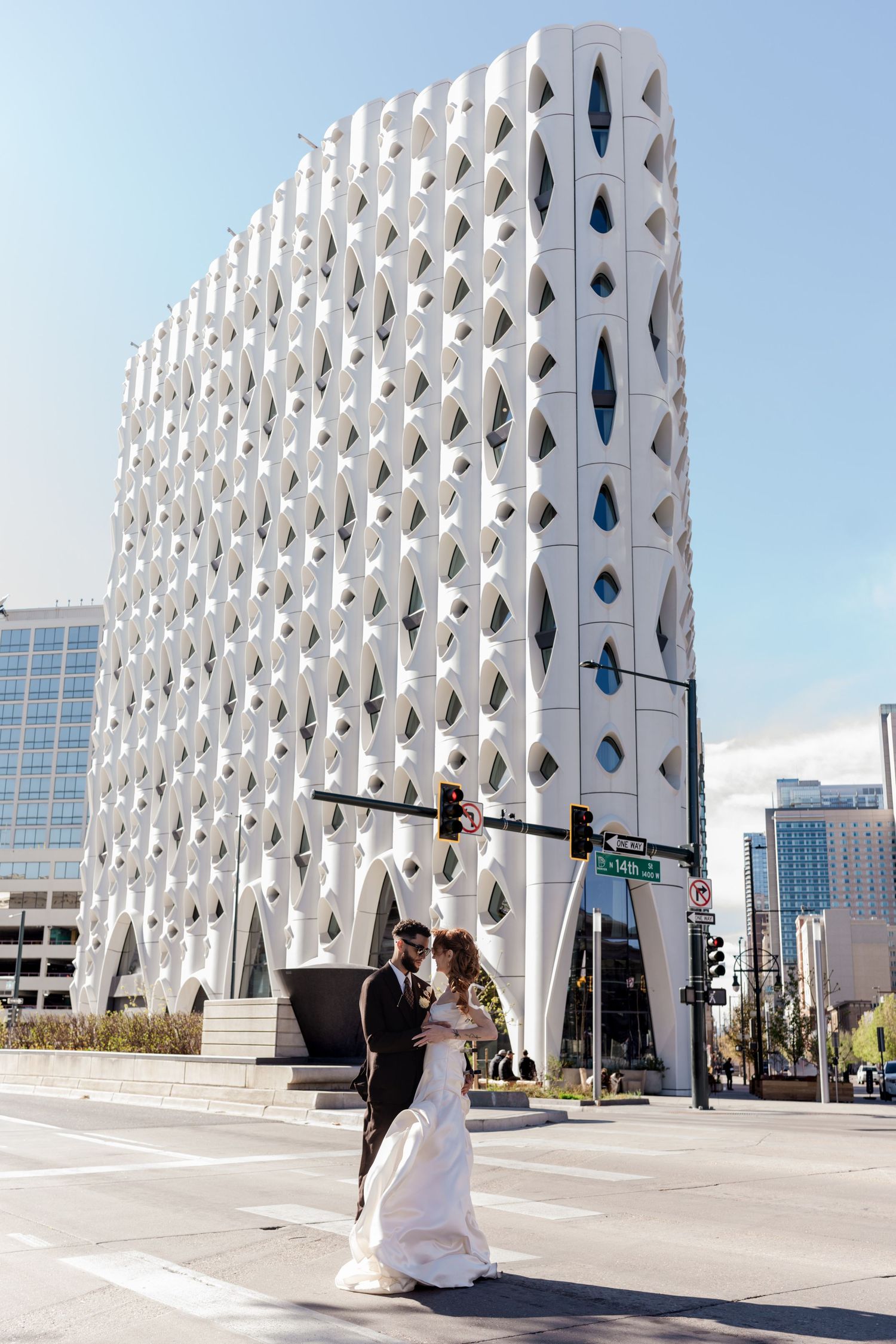 Bride and groom pose at urban intersection with distinctive white geometric building facade in city background.