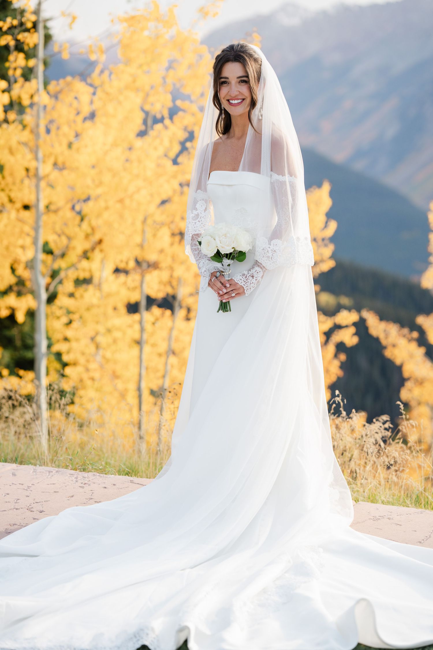 Bride in elegant white gown with veil holding white bouquet surrounded by golden autumn foliage.
