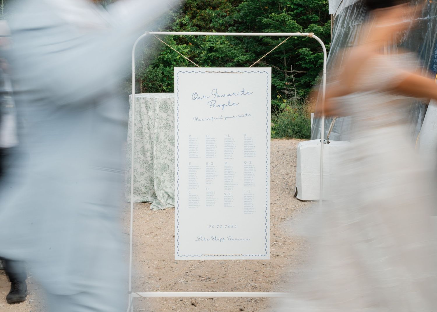Blurred guests walk past a white framed wedding seating chart sign outdoors.