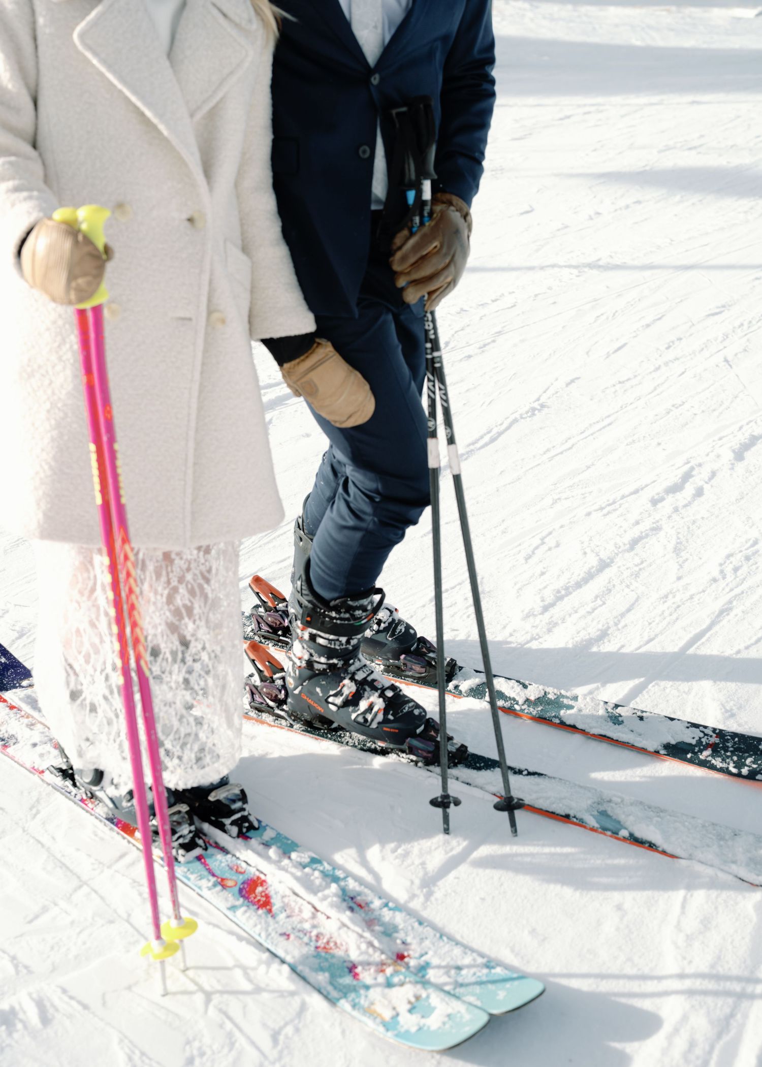 Close-up of bride in lace skirt and groom in navy suit standing on skis in fresh snow.