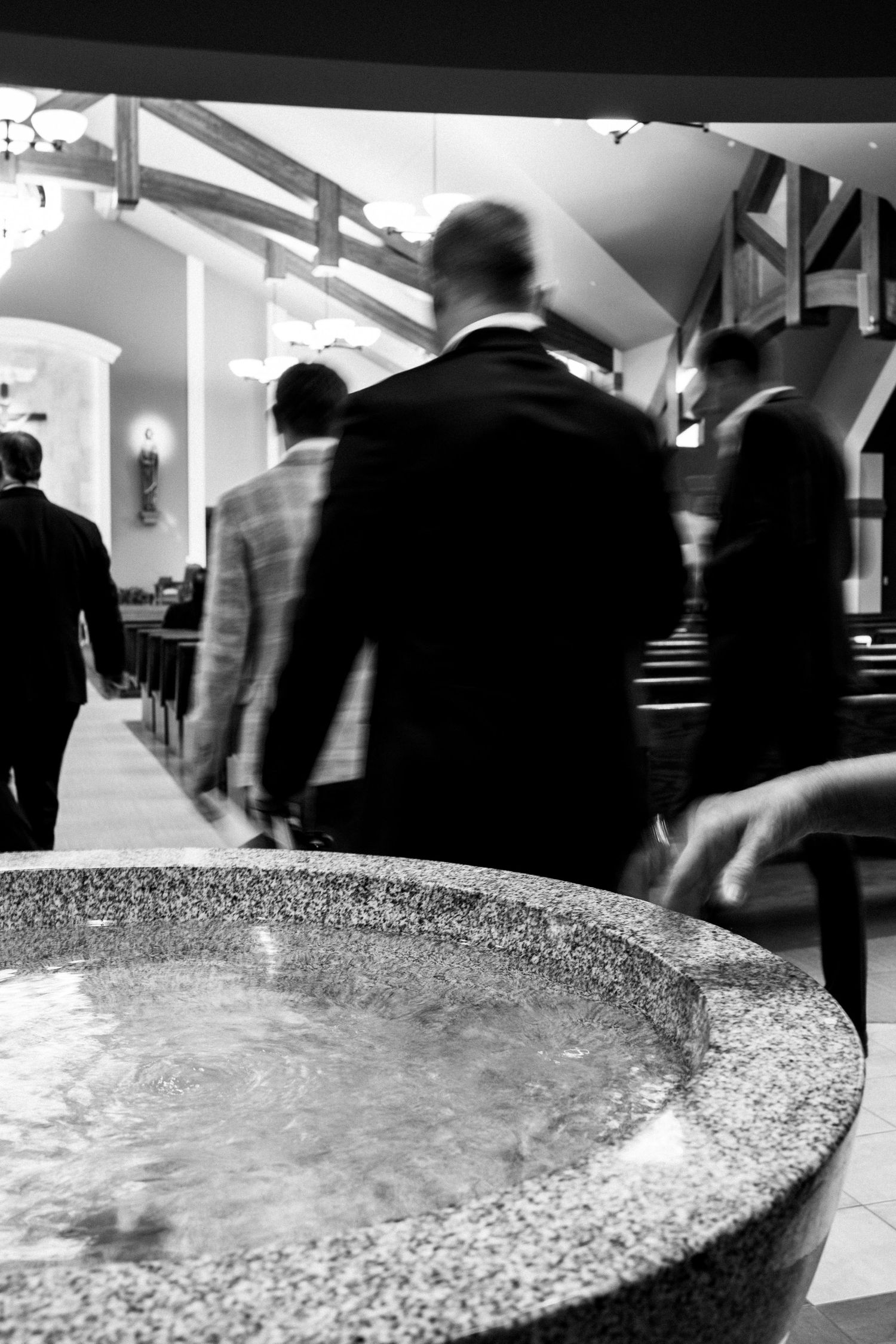 Black and white photo of groomsmen walking past holy water font inside a church before wedding ceremony.