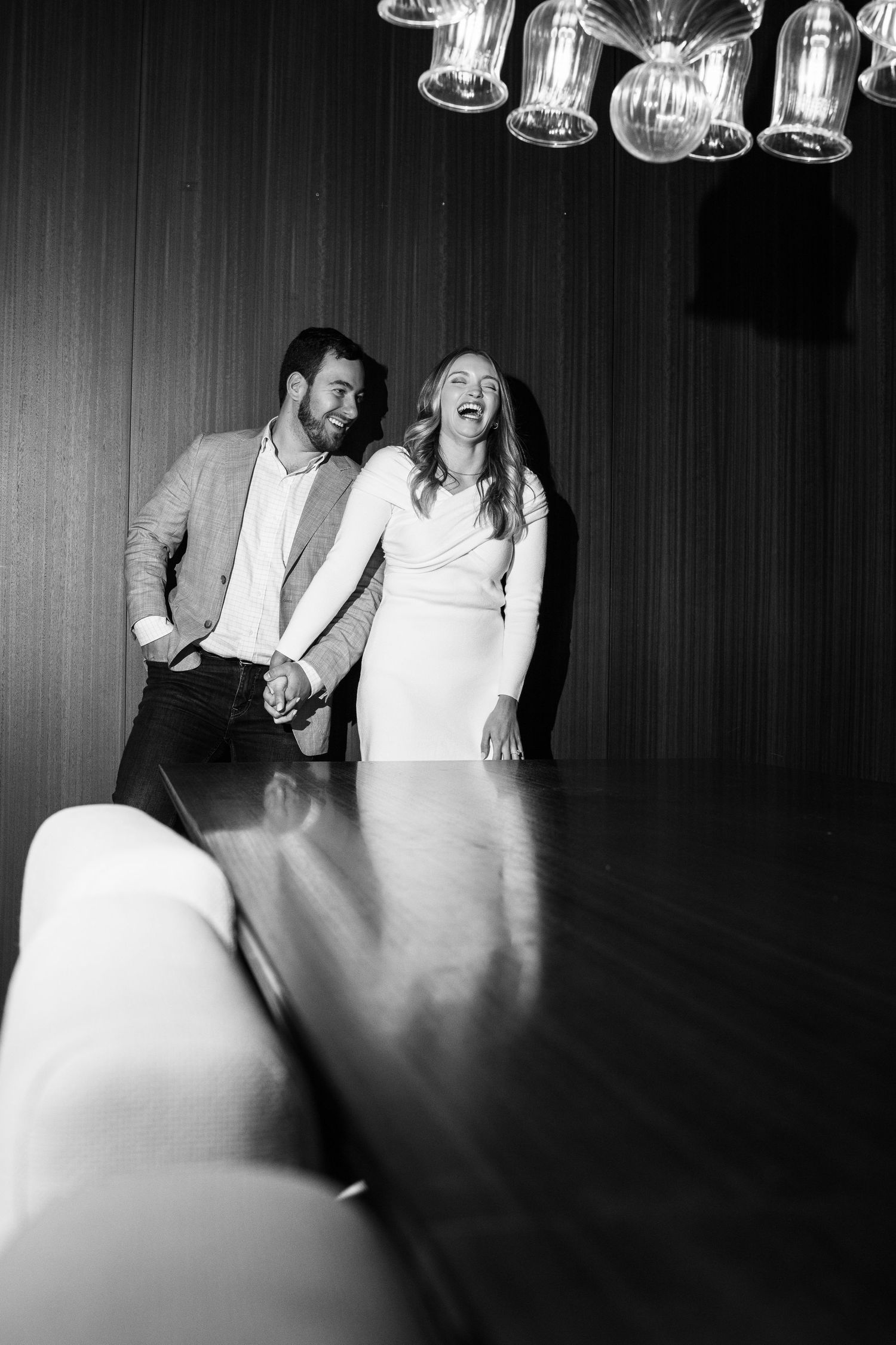Black and white photo of joyful couple laughing together under elegant chandelier lights in stylish venue.