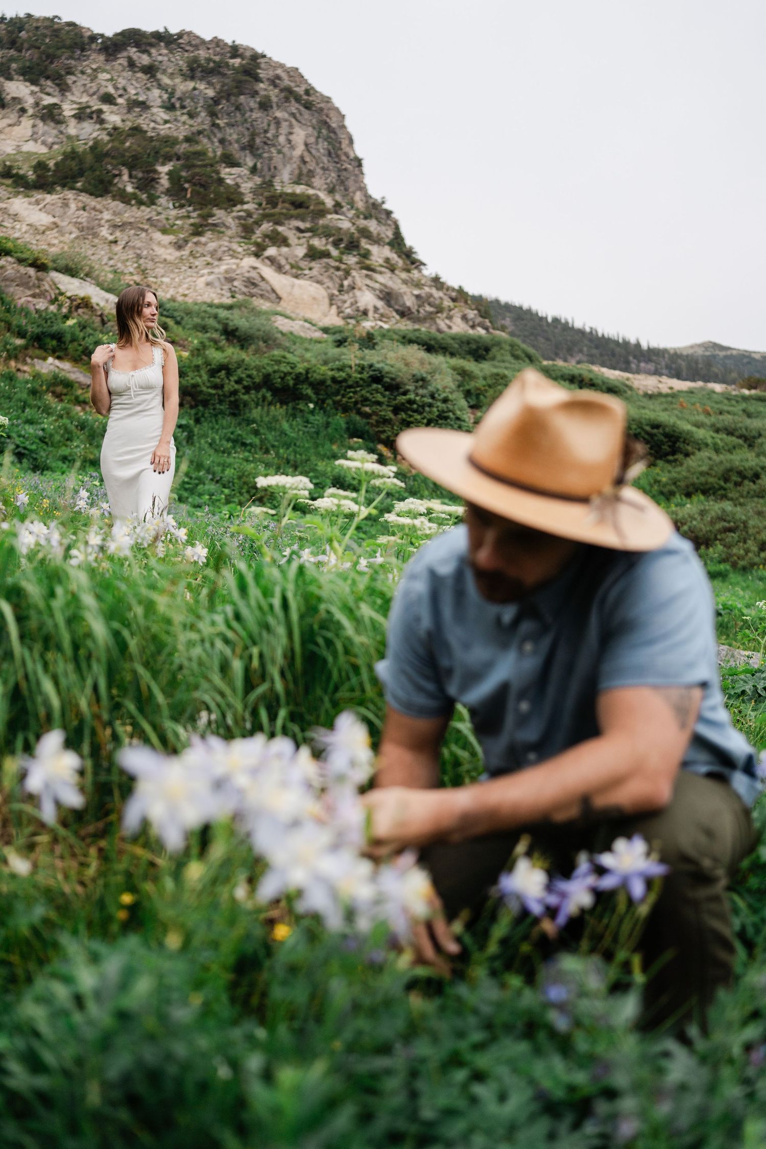 Woman in white dress walking through wildflower meadow toward man picking flowers near rocky mountain backdrop.