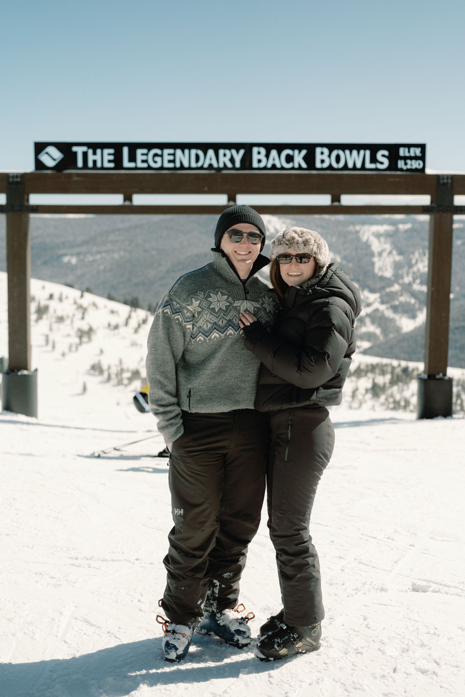 A happy couple poses together at The Legendary Back Bowls ski resort sign on a bright sunny winter day.