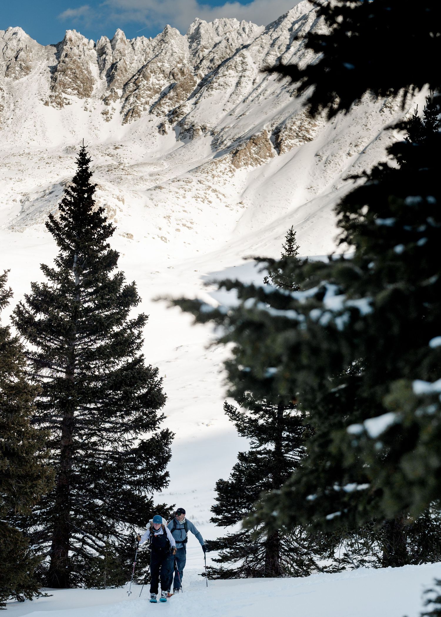 Two skiers climb a snowy trail framed by tall pine trees with rugged snow-covered mountain peaks behind them.