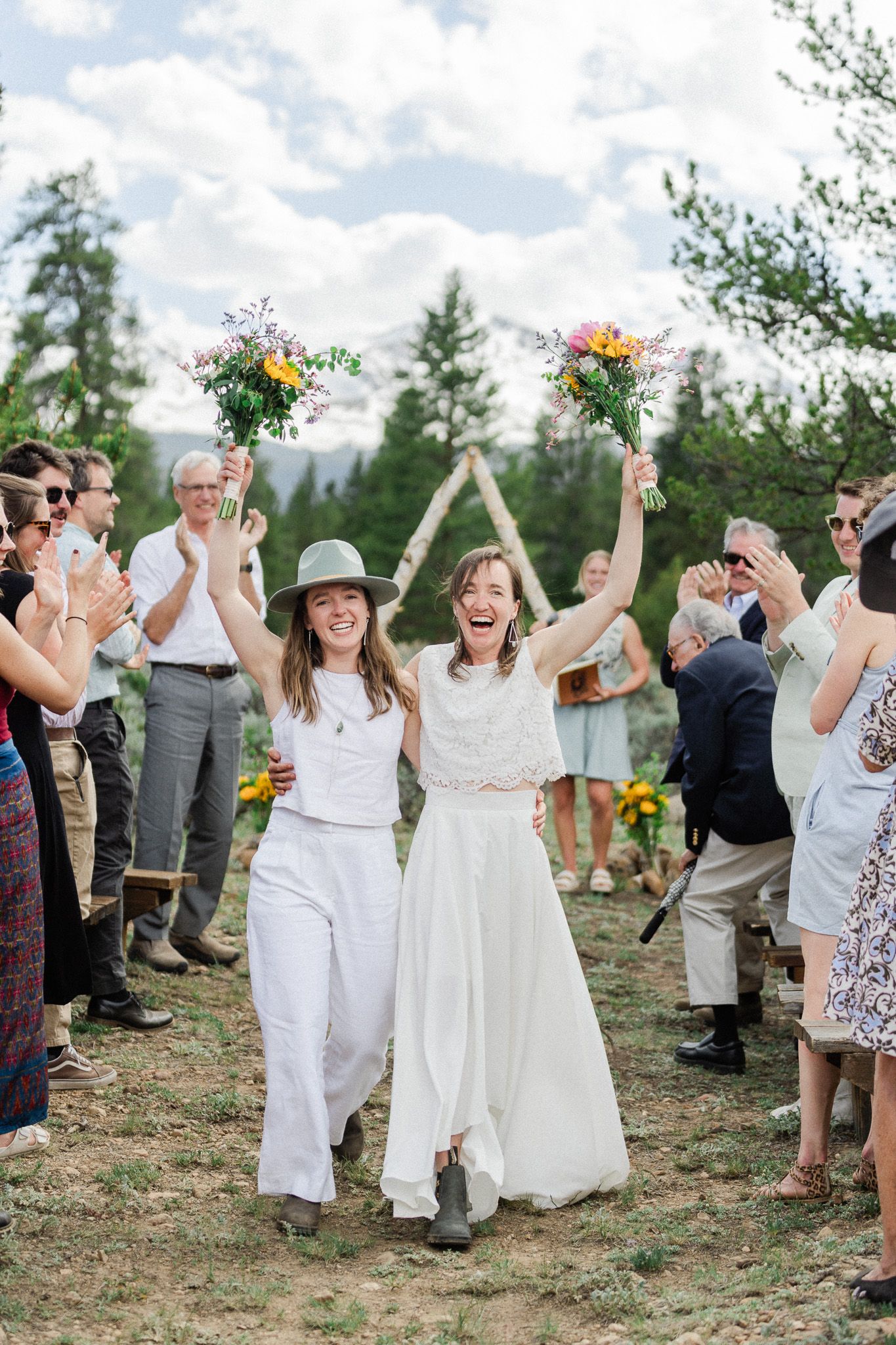 Two brides celebrating joyfully with raised bouquets as guests cheer at outdoor mountain wedding.