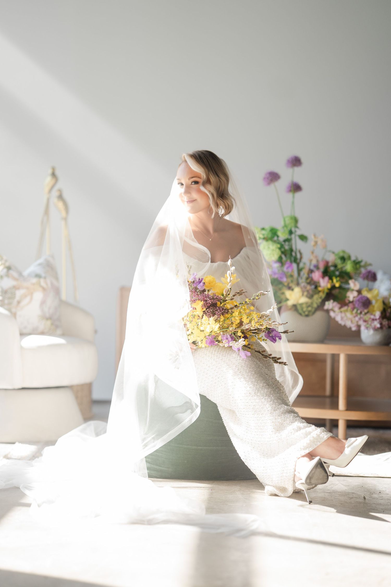 Bride in white gown and veil sitting in sunlit room holding colorful yellow and purple bouquet.