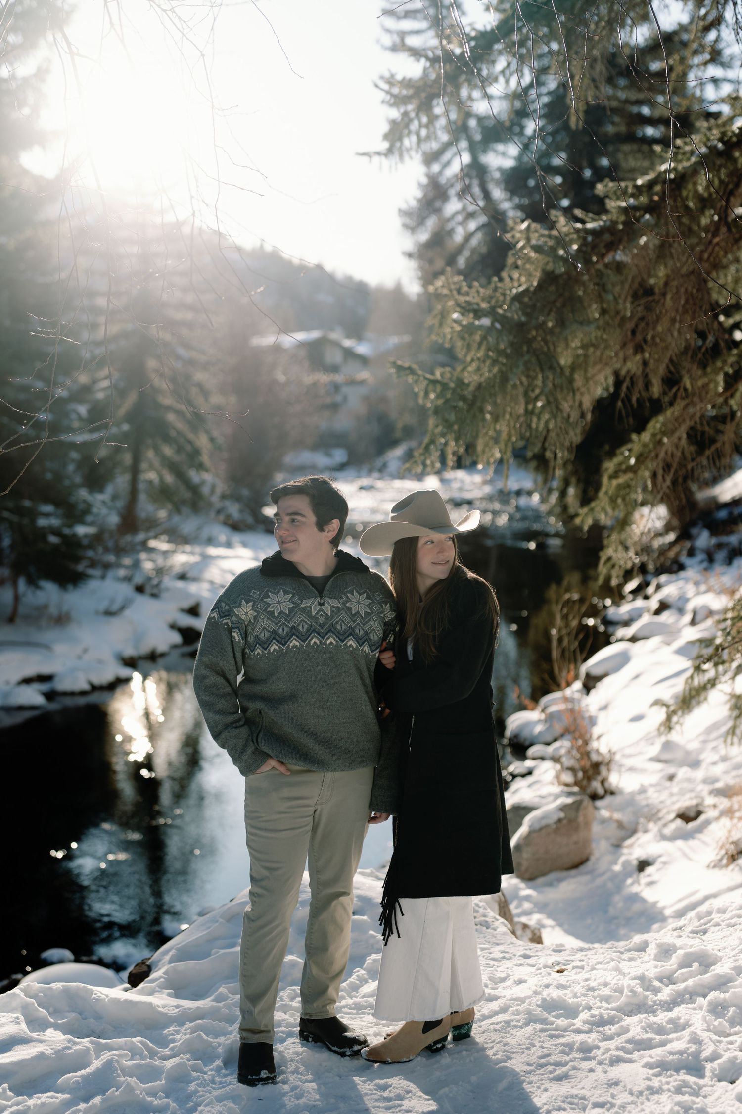 Couple standing together in snowy forest by a creek with golden sunlight filtering through trees.
