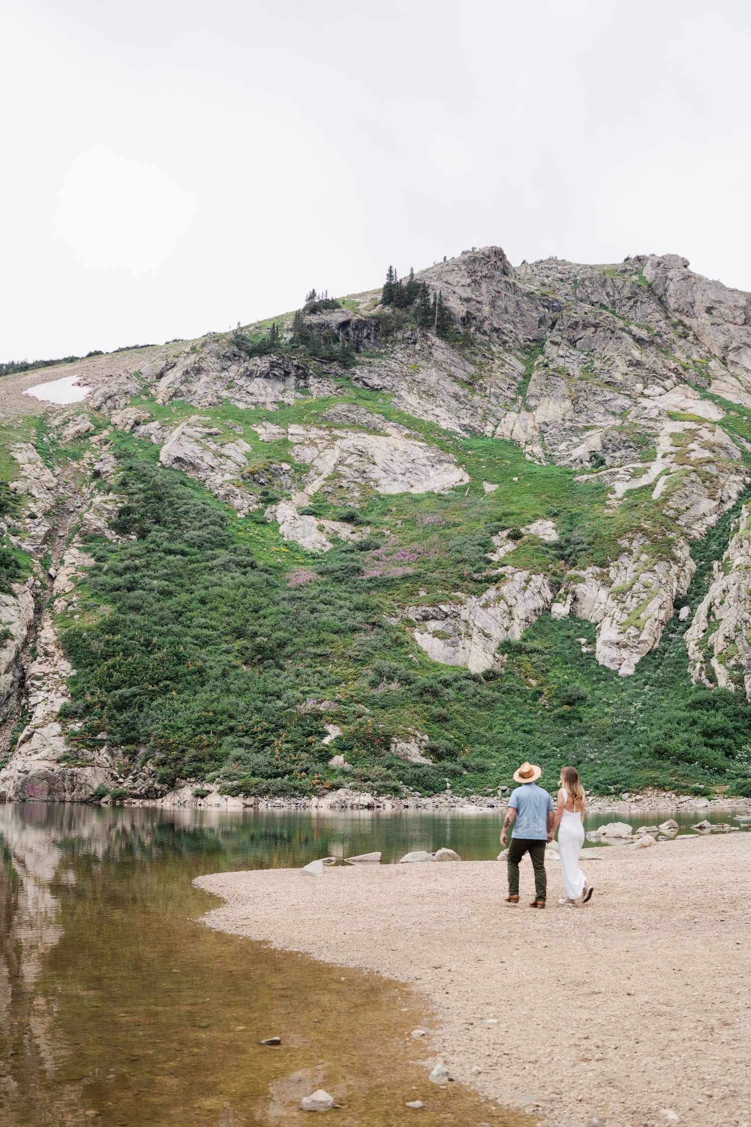 Couple holding hands walking along rocky alpine lake shore with dramatic mountain backdrop.