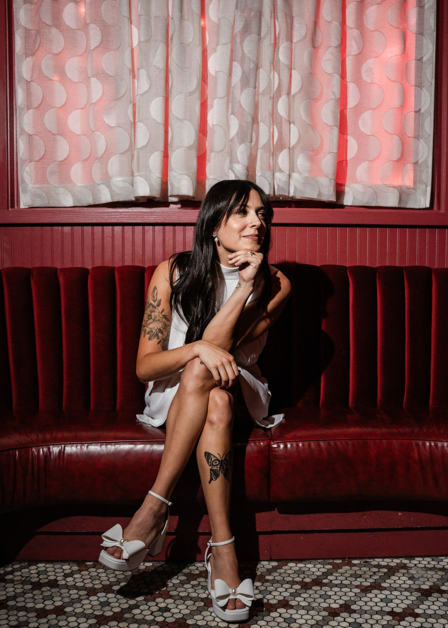 Tattooed woman in a white mini dress and platform heels seated on a red velvet lounge sofa.