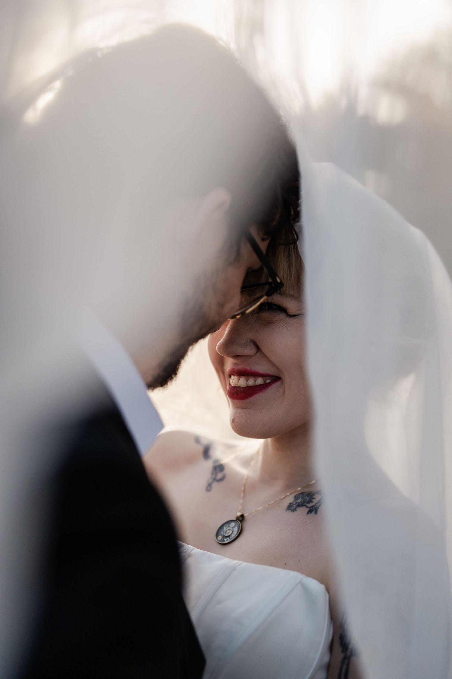 Smiling tattooed bride with red lips and vintage necklace peeking through flowing white wedding veil.