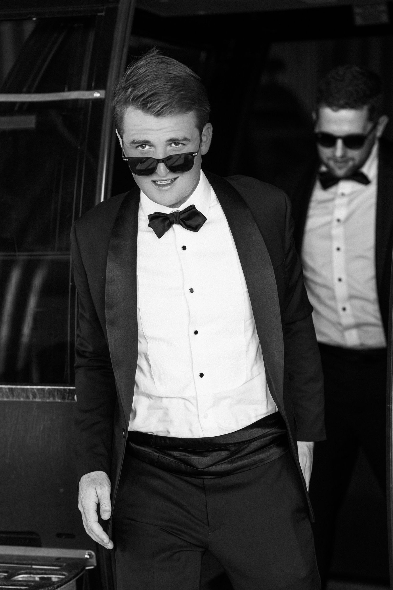 Stylish young man in black tuxedo and sunglasses smiling at a formal event in black and white.