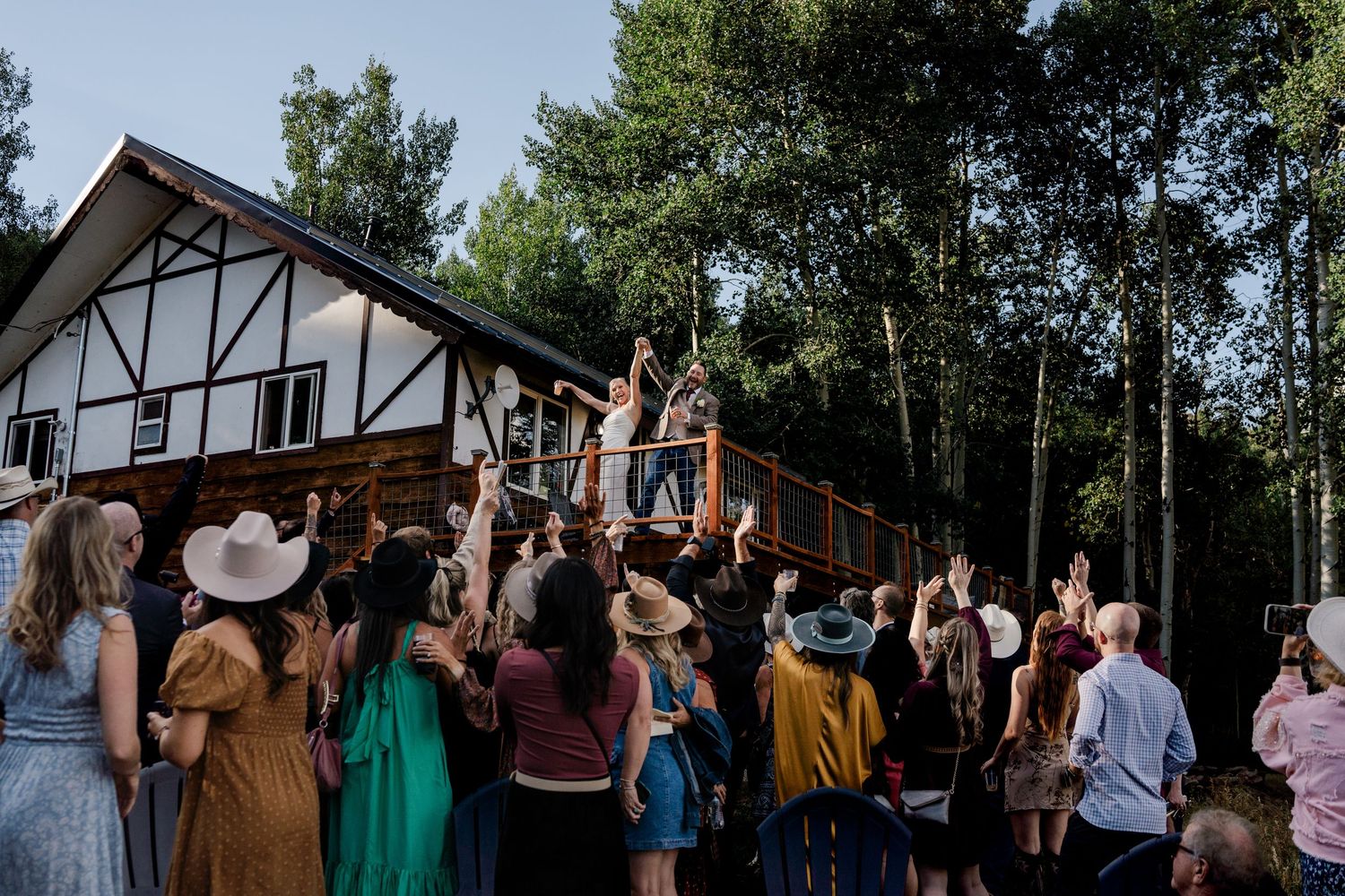 Wedding guests cheering as bride and groom celebrate on a rustic wooden deck surrounded by tall aspen trees.