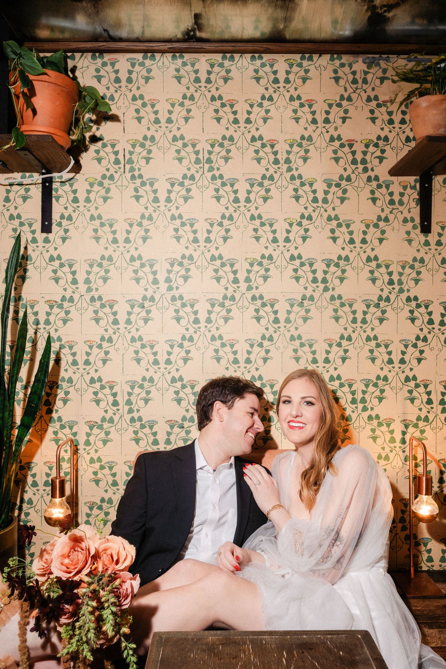 Couple sitting close together in a cozy booth with vintage green floral wallpaper and warm lighting.