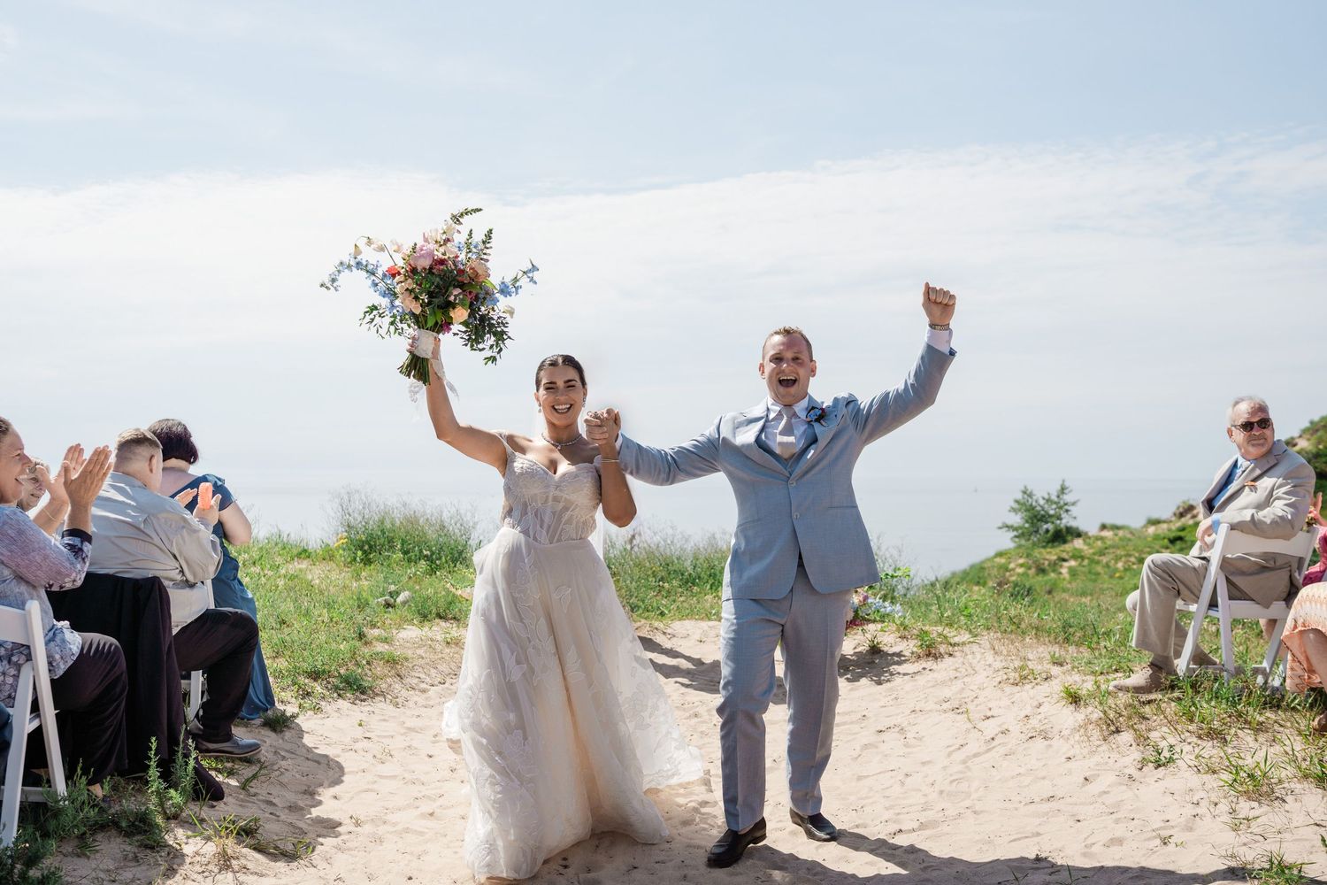 Joyful newlyweds celebrate at sandy outdoor ceremony, bride holding colorful bouquet aloft under blue sky.