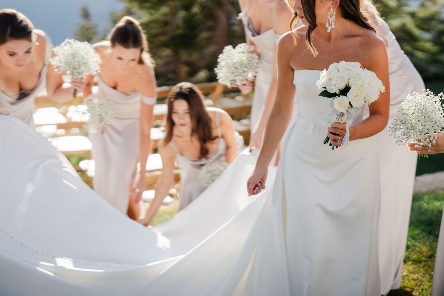 Bridesmaids in champagne gowns help adjust the bride's white wedding dress train at an outdoor mountain venue.
