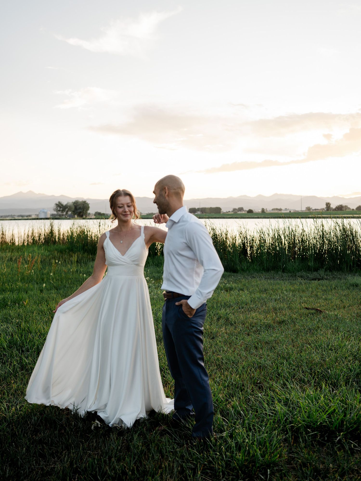 A bride in a white gown and groom in navy suit share a romantic moment by a lake at sunset with mountain views.
