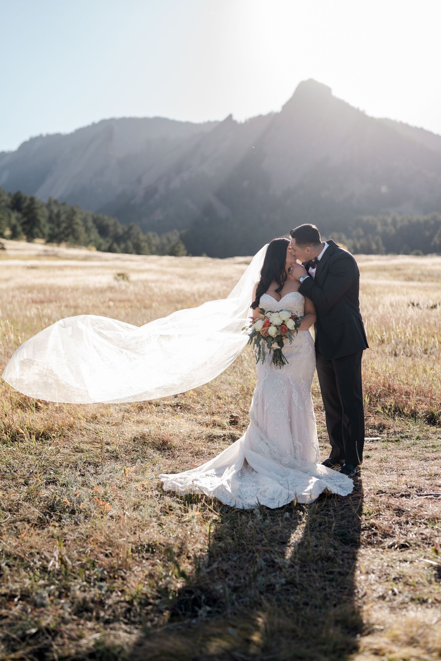 Bride and groom kissing in sunlit field as wedding veil billows in wind with Flatirons behind.