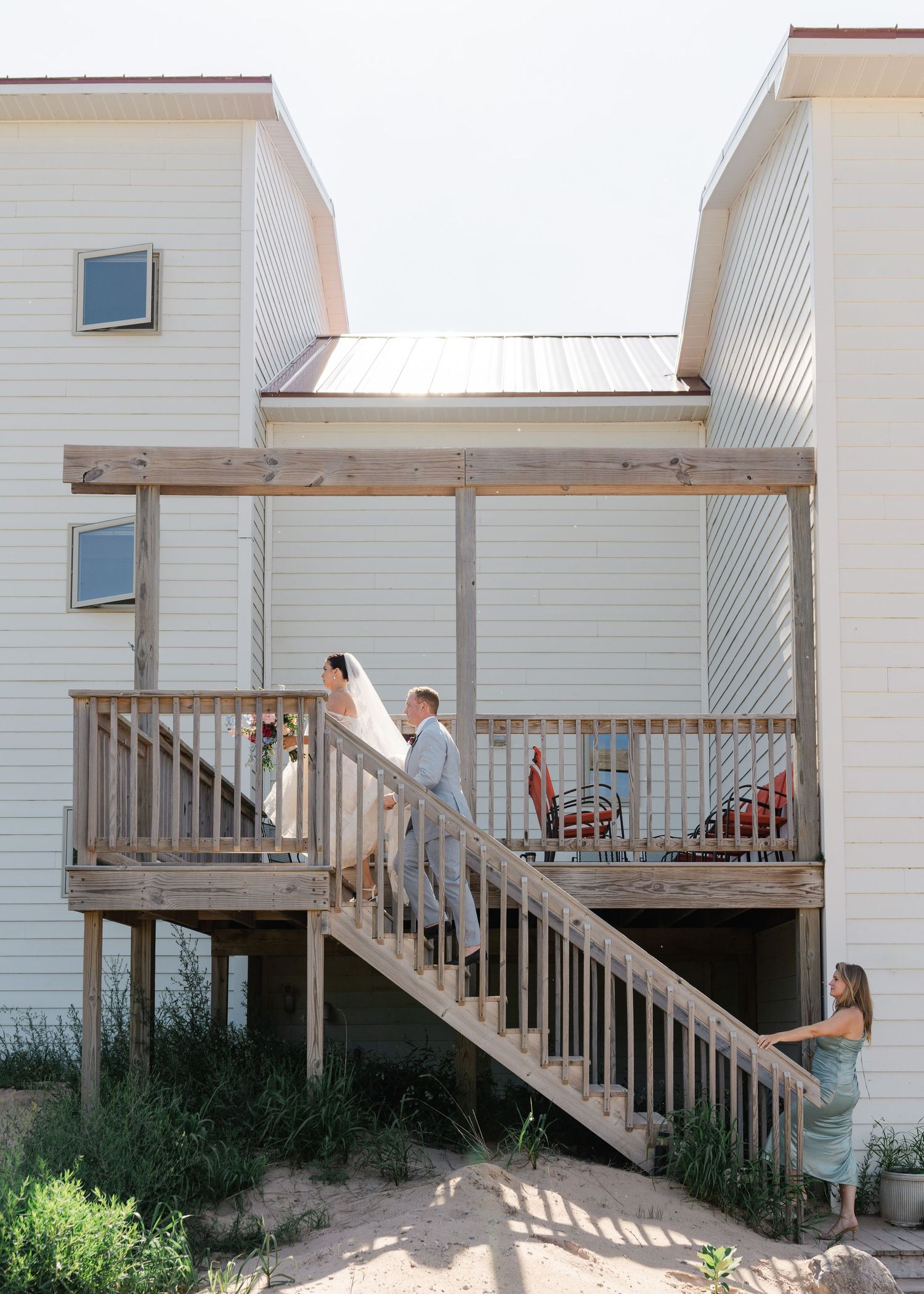Bride and groom walk up wooden staircase of coastal wedding venue while bridesmaid watches from below.