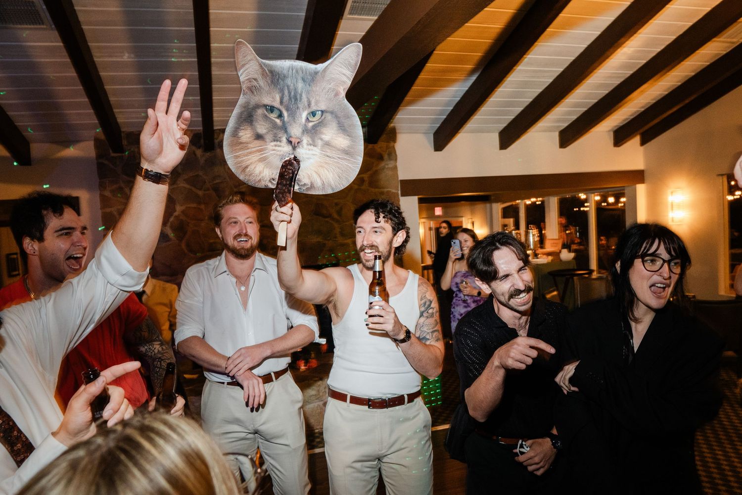 Group of men laughing and holding a large cat face cutout at a lively wedding reception party.