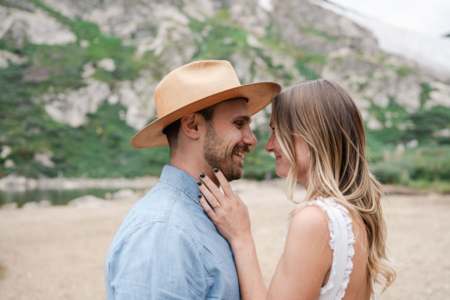 Smiling couple in tender moment, man in tan hat and blue shirt with mountain backdrop.