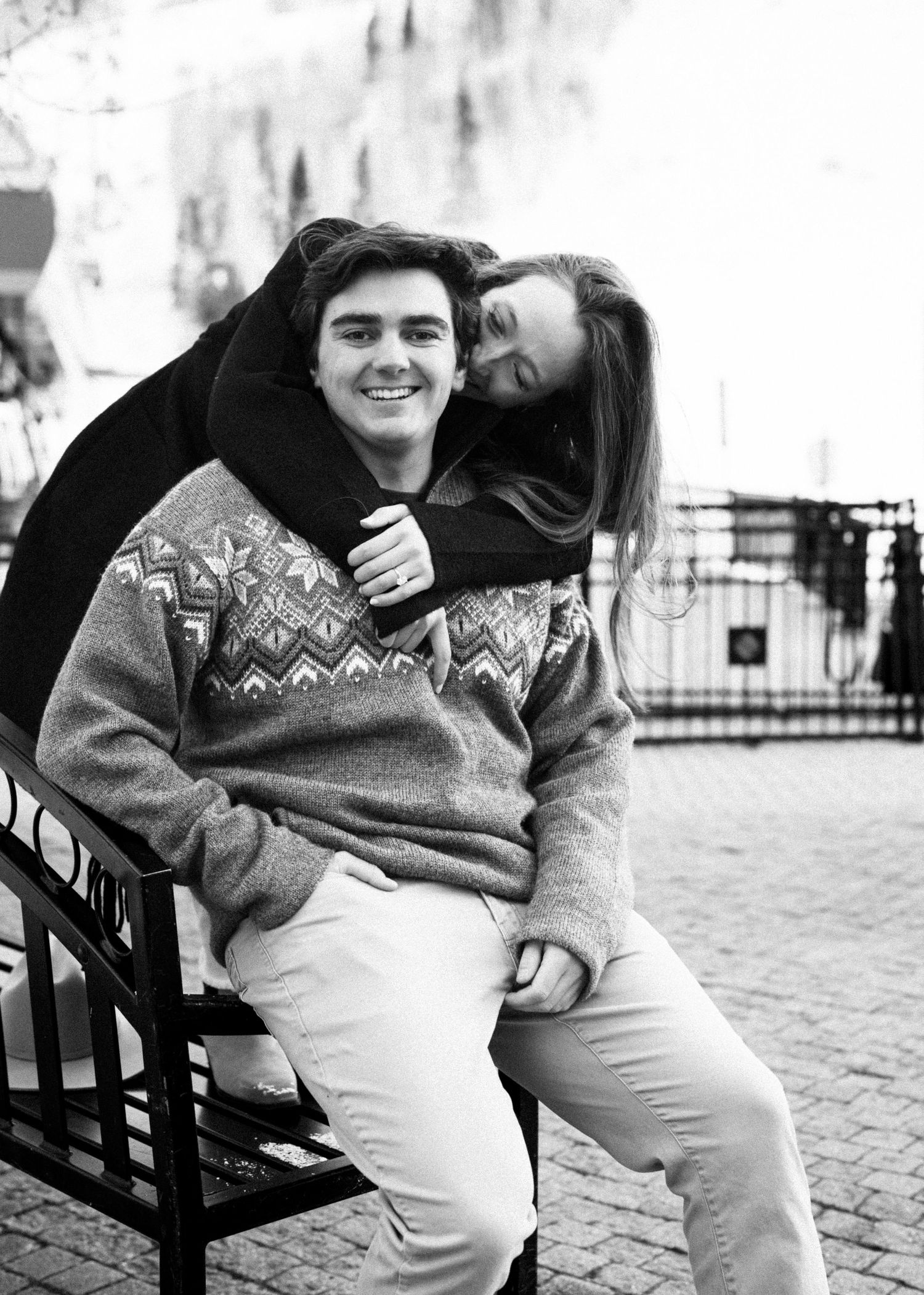 Black and white photo of a young couple laughing on a bench, man in a Nordic sweater and khakis.