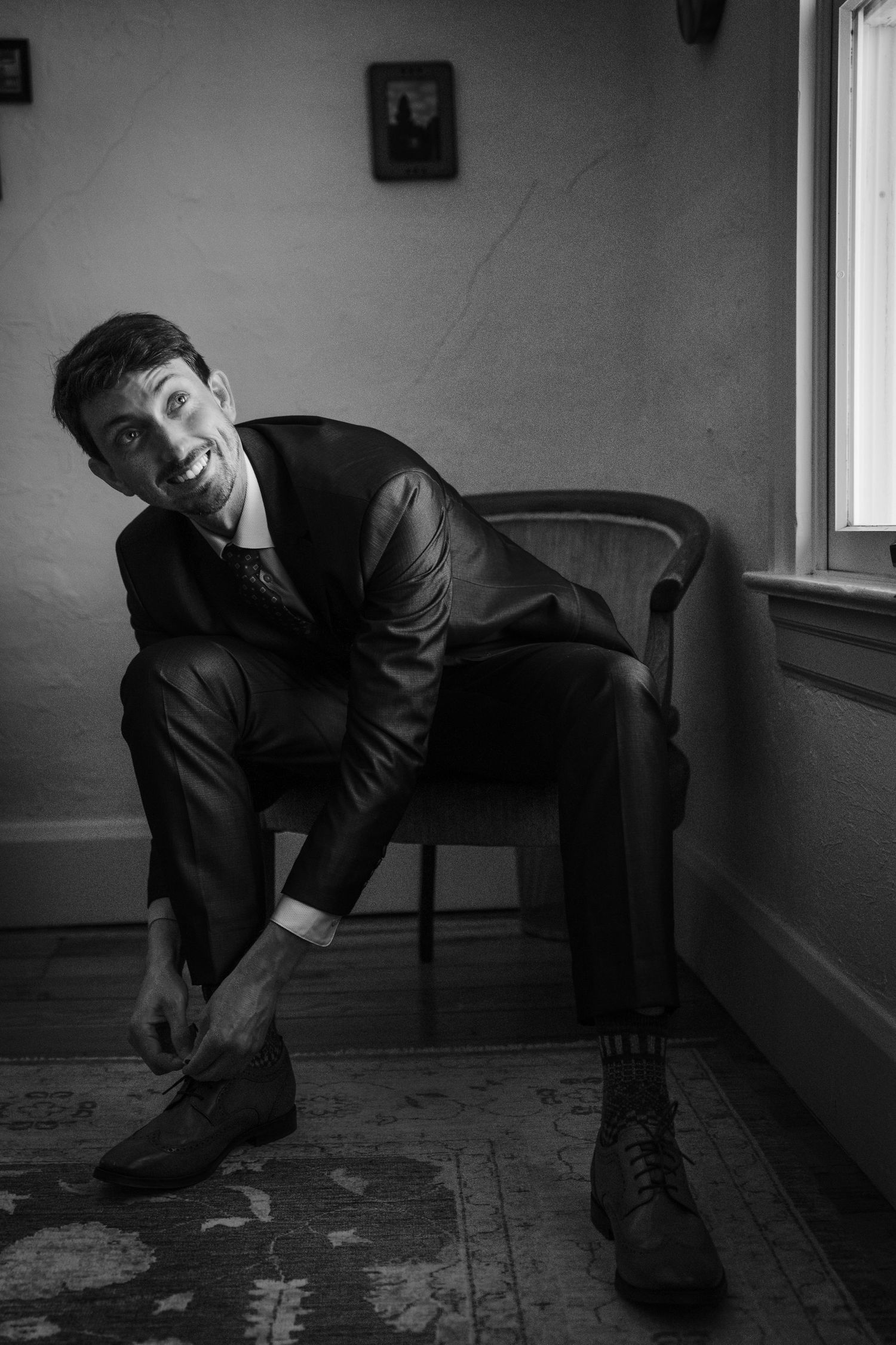 Groom smiling while putting on dress shoes in a chair, wearing a black suit in a black and white photo.