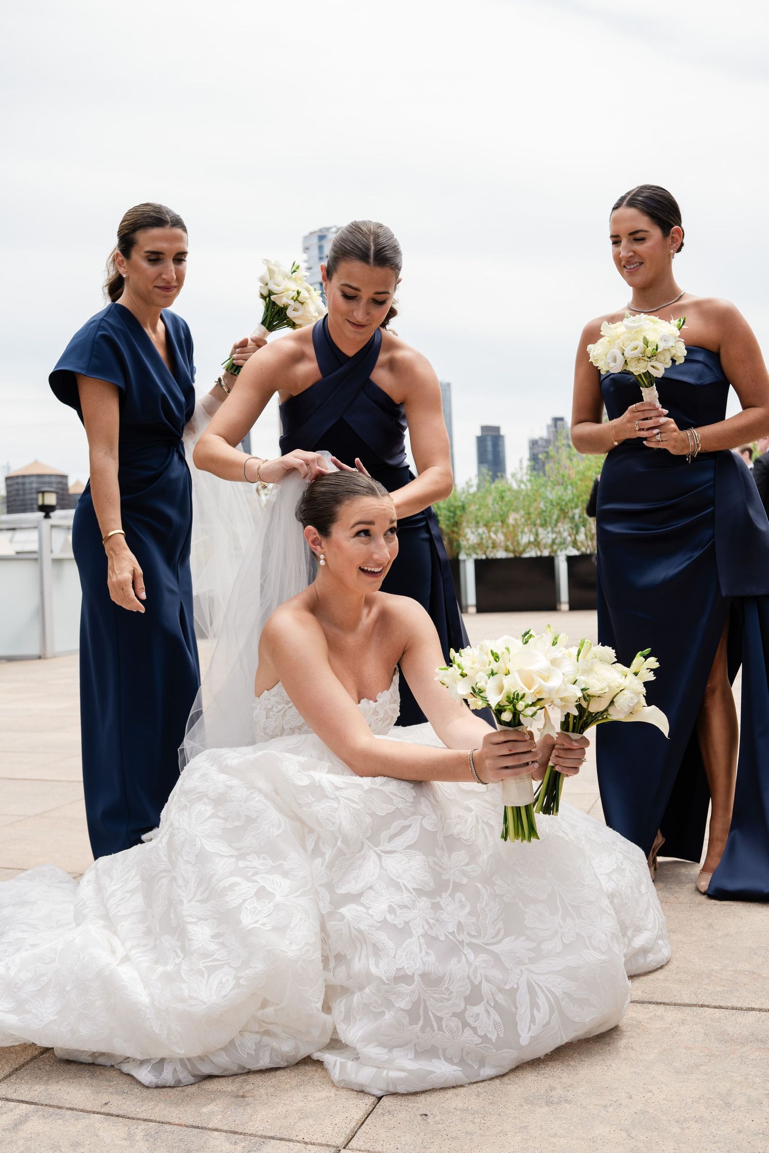 Bride in lace ball gown sitting as bridesmaids in navy dresses adjust her veil on outdoor terrace.