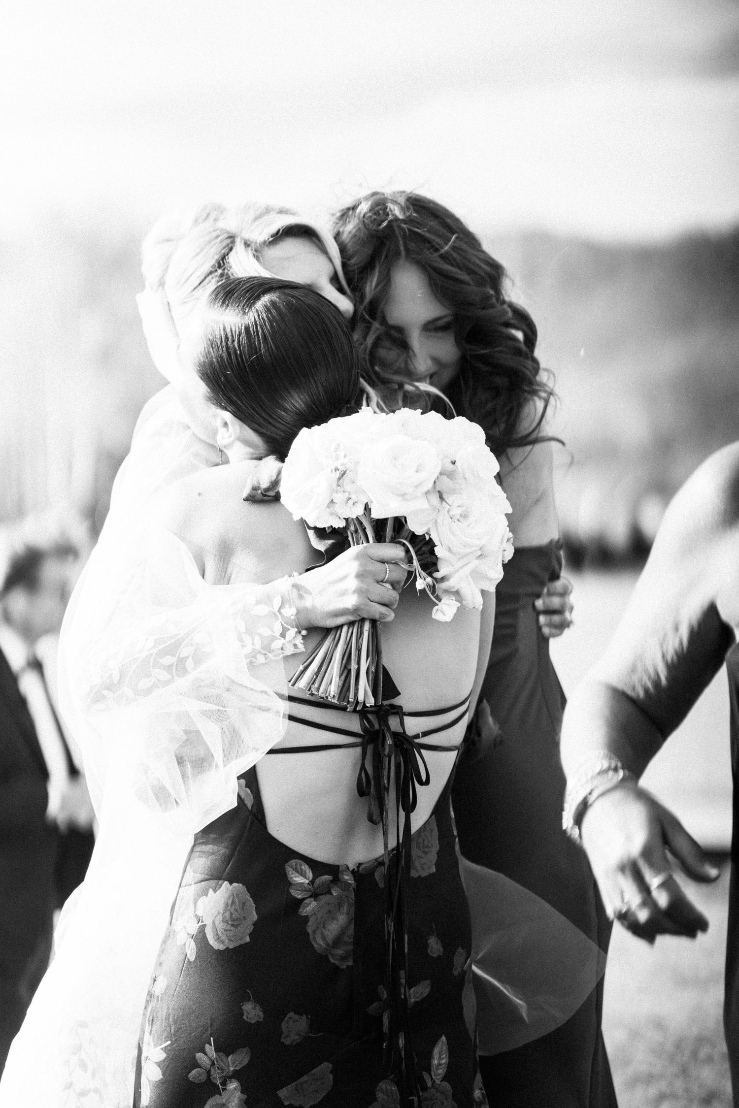 Black and white photo of bride embracing bridesmaid in floral dress while holding white rose bouquet at wedding.