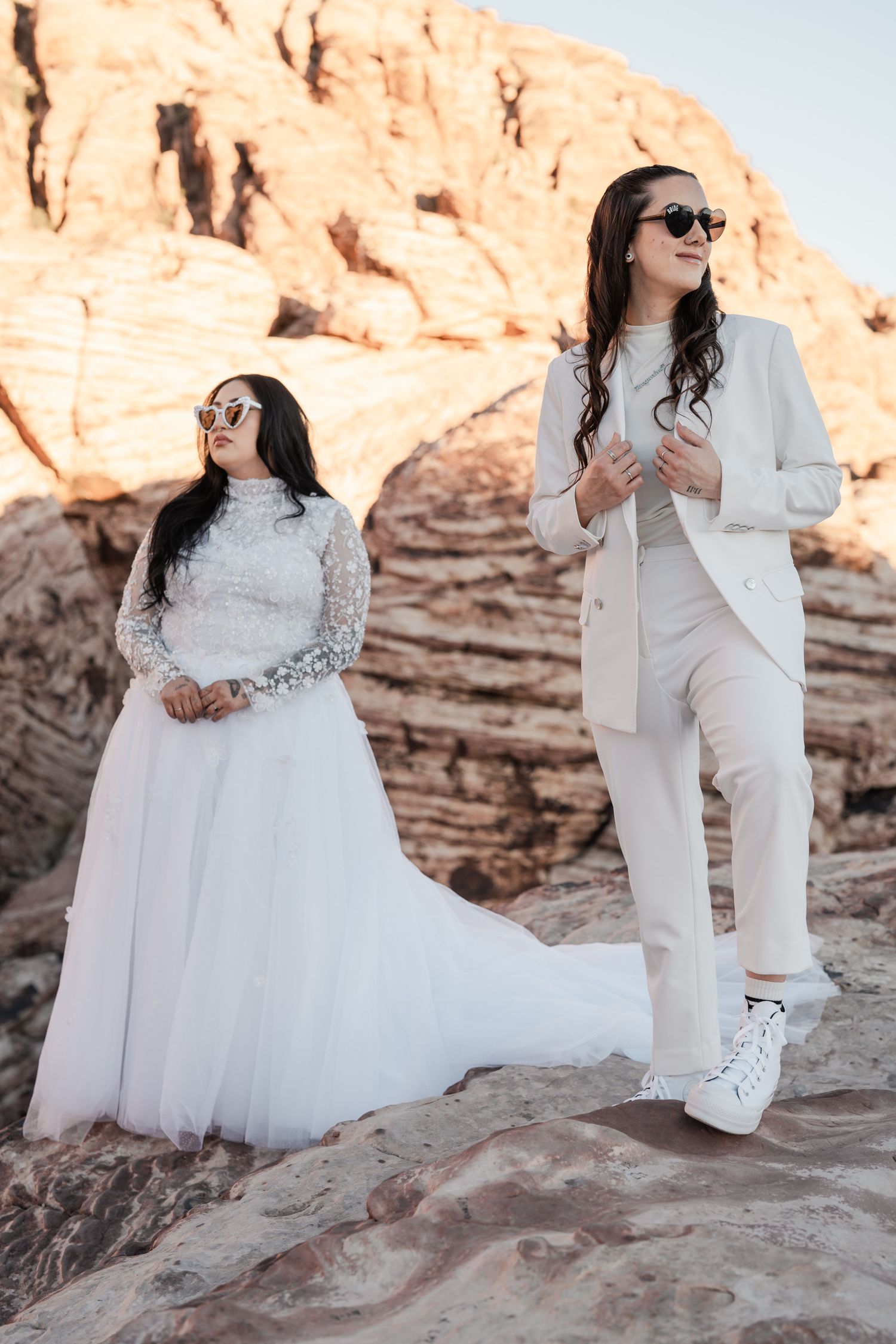 Two brides in white wedding attire pose on desert rocks at Red Rock Canyon at sunset.