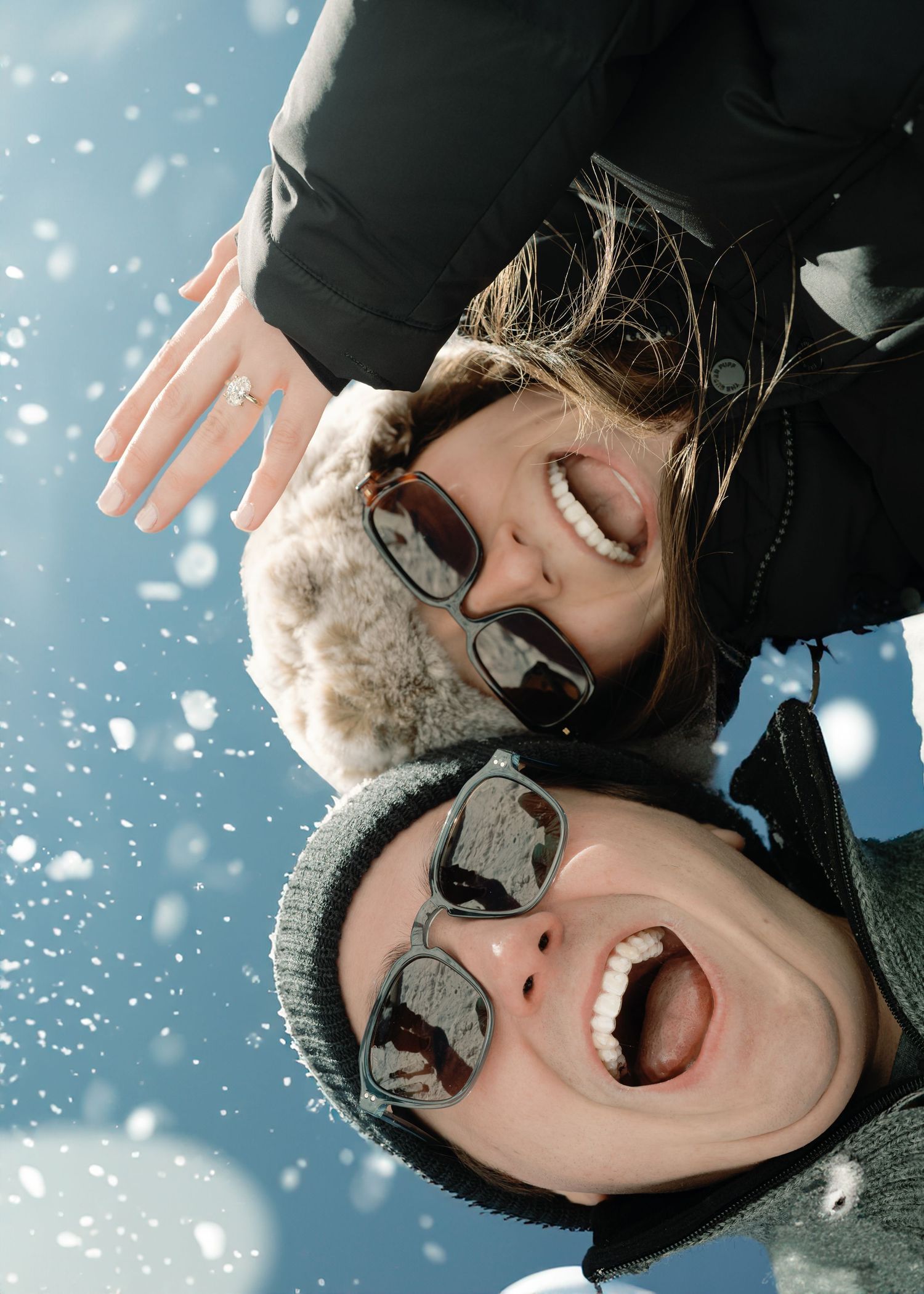 A joyful couple laughs upside down together in the snow, one showing off an engagement ring on a sunny winter day.