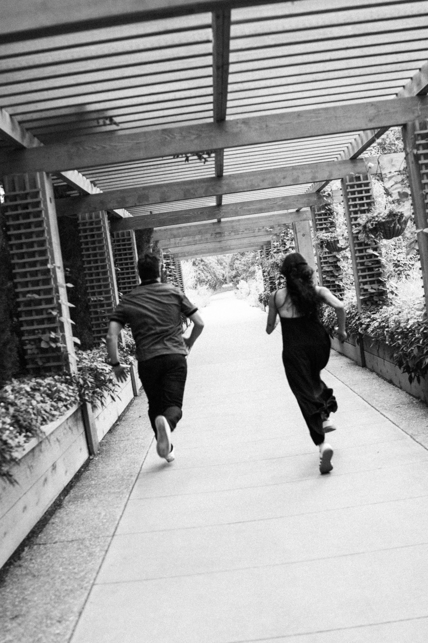 Black and white photo of couple running hand-in-hand through pergola walkway in garden.