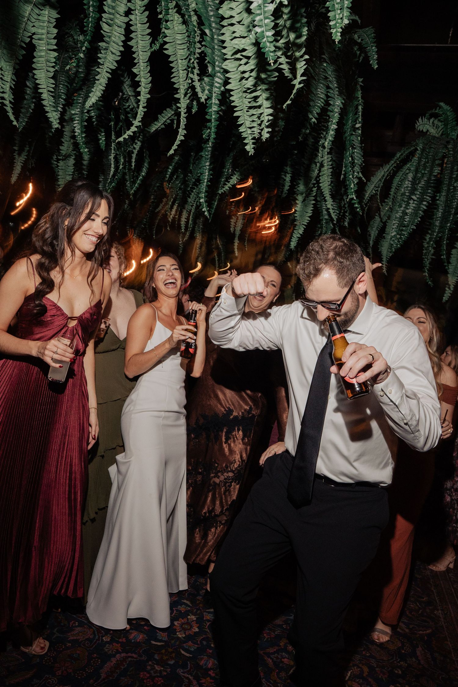Wedding guests dancing energetically under hanging ferns, groom in white shirt and tie holding beer bottle.