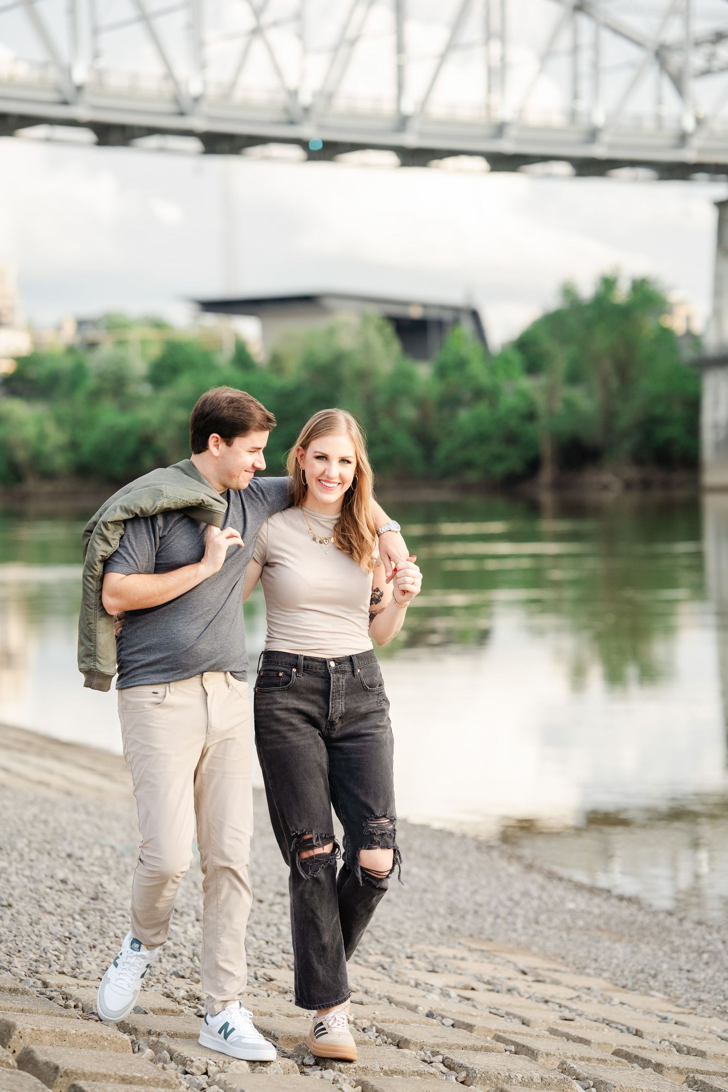 Happy couple walking along a riverbank with a bridge and green trees in the background.