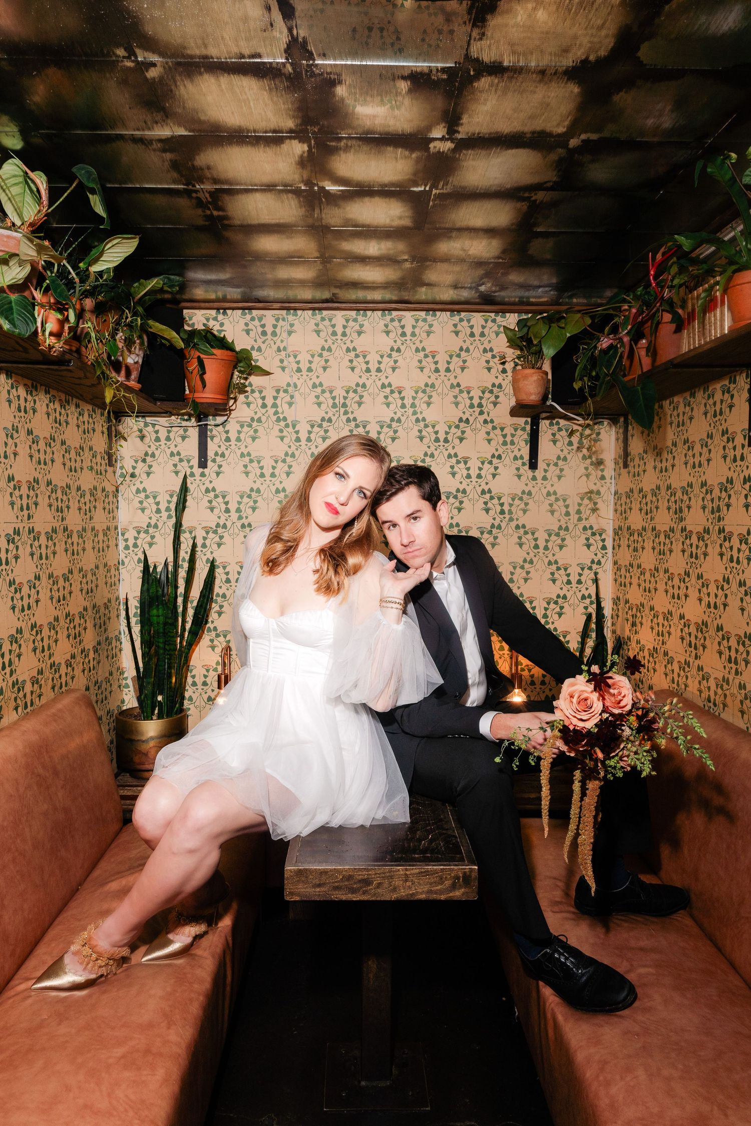 Bride and groom pose together in a cozy botanical booth with green wallpaper and hanging plants.
