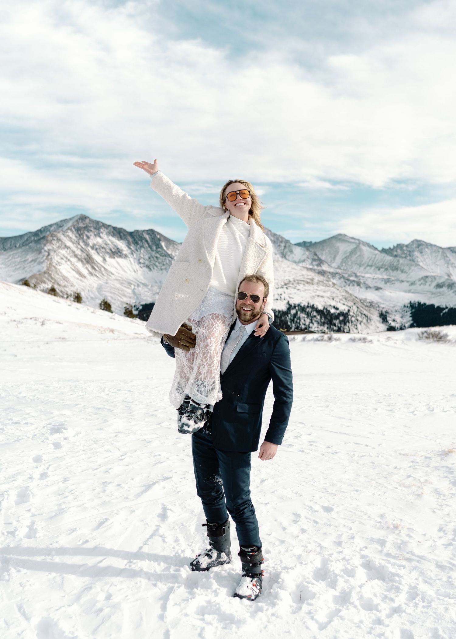 Couple celebrating winter wedding in snowy mountain landscape, bride lifted on groom's shoulders in white dress.