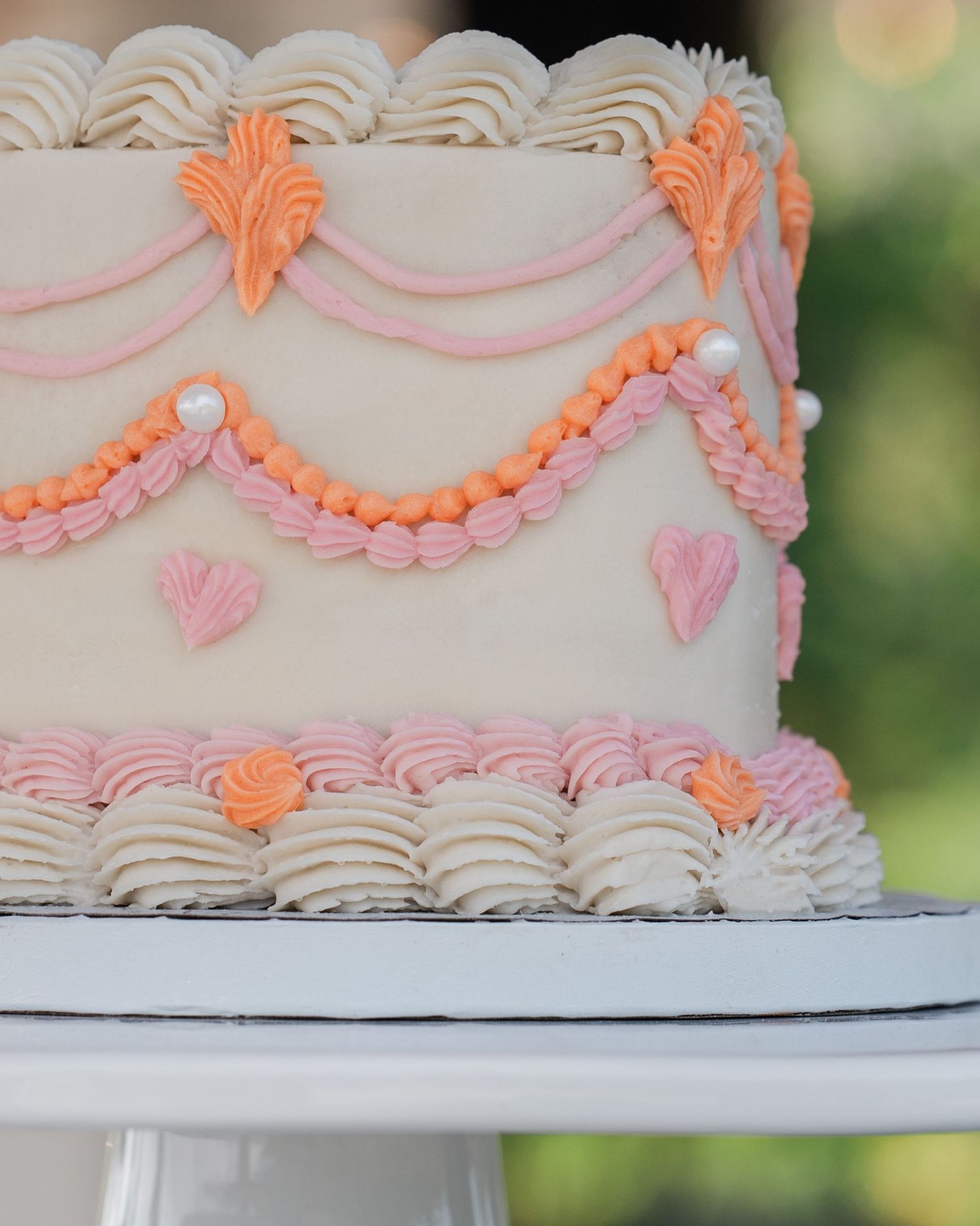 Elegant two-tier wedding cake decorated with pink and orange buttercream swags, hearts, and pearls.