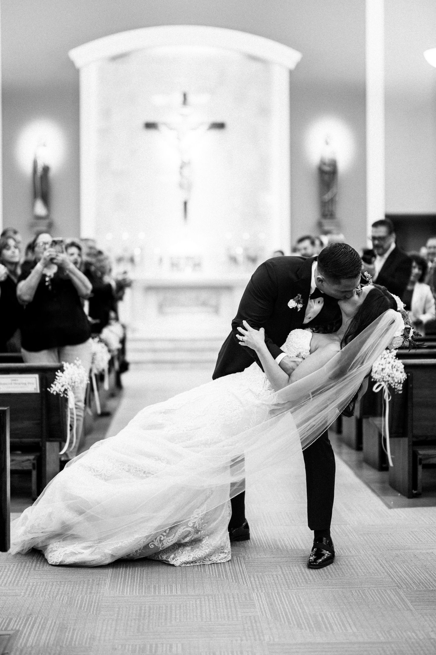 Groom dipping bride for romantic kiss in church aisle after wedding ceremony in black and white.