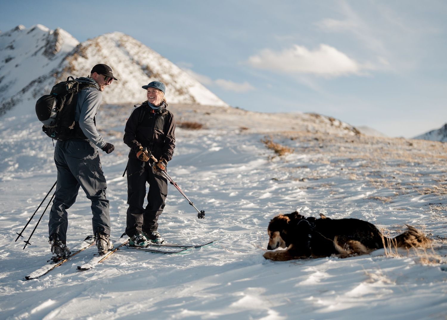 Two backcountry skiers pause on a snowy slope to check gear while their dog rests in the snow nearby.