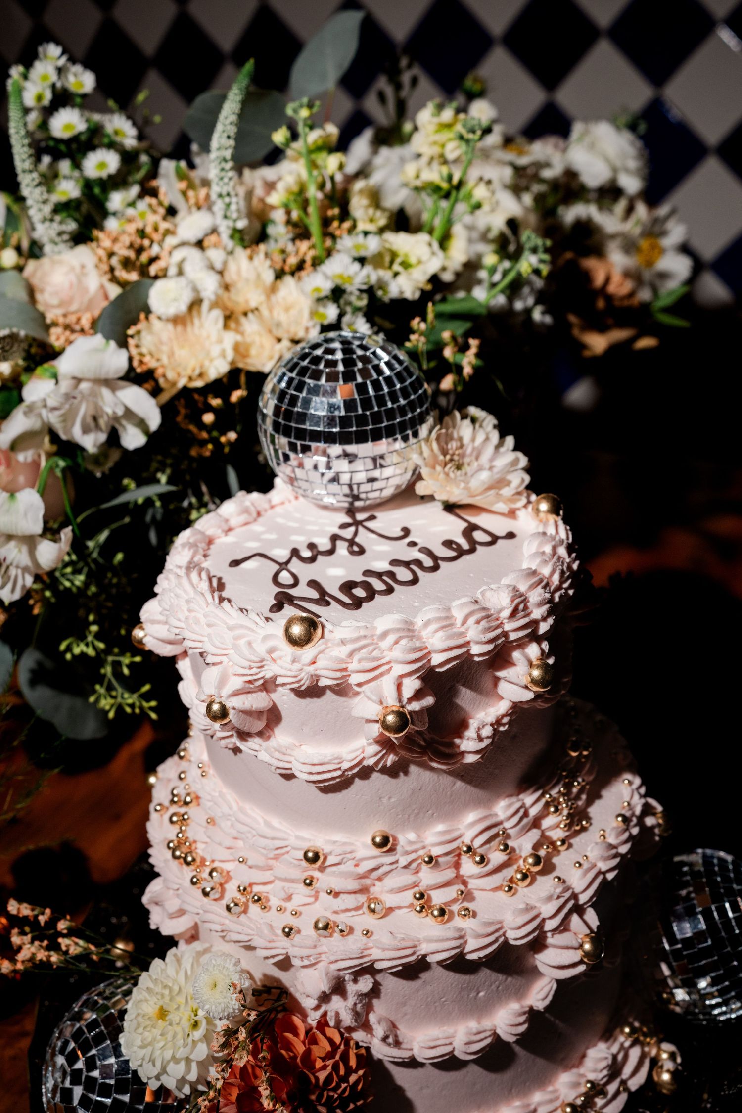 Pink three-tier Just Married wedding cake with disco ball topper and gold pearls surrounded by floral arrangement.