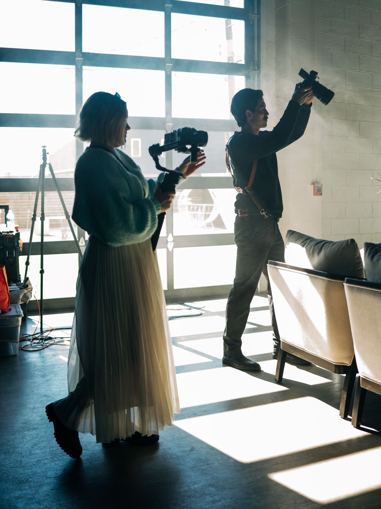 Two videographers holding cameras in a sunlit studio space with chairs and a tripod in the background.