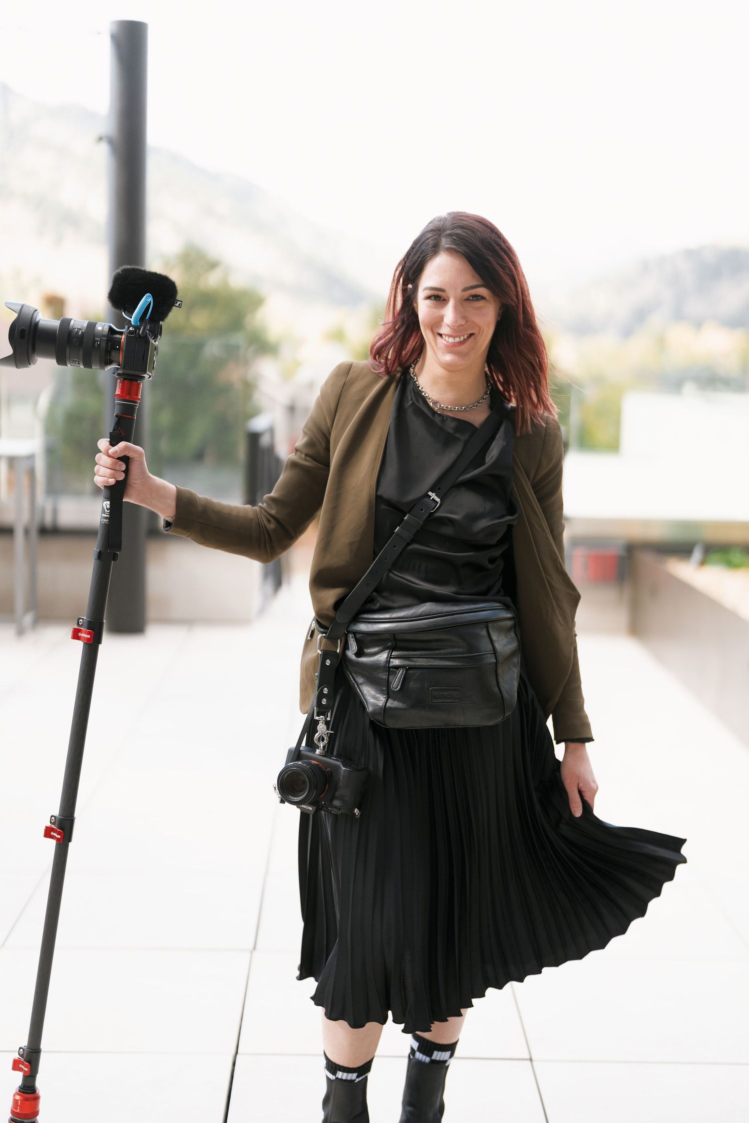 Smiling female photographer in black pleated skirt and olive blazer holding a camera monopod outdoors.