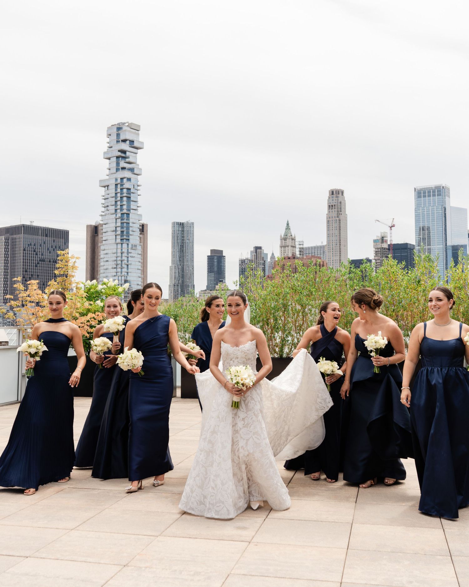 Bride in lace gown with bridesmaids in navy dresses on rooftop with NYC skyline backdrop.