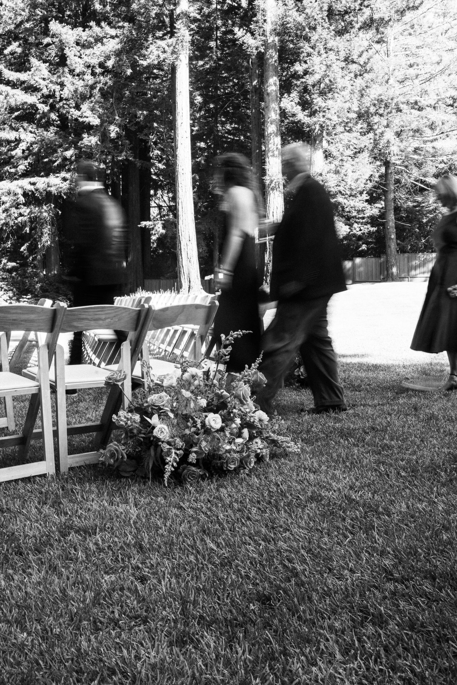 Black and white photo of wedding guests walking past floral arrangement on grass aisle.